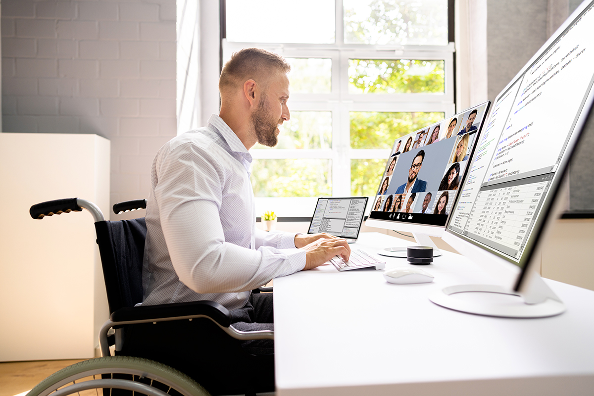 homme en fauteuil roulant travaille devant son ordinateur avec deux ecrans dans son bureau