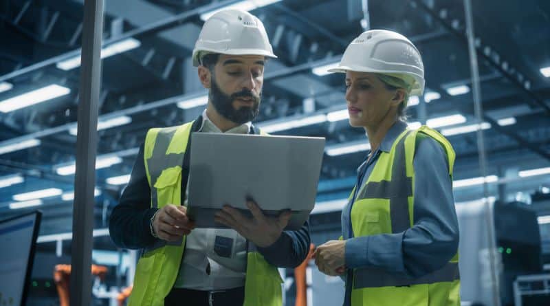 un homme et une femme sont au premier plan. ils portent un casque de chantier et un gilet. Ils échangent en regardant un ordinateur portable. A l'arrière-plan de trouve un espace technique.