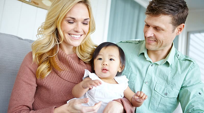 Un couple européen souriant assis sur un canape, la femme a une petite fille asiatique sur les genoux.