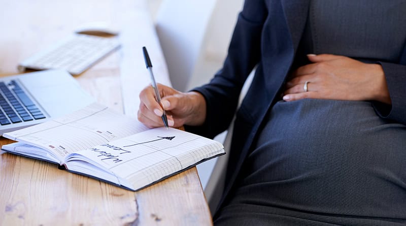 Femme enceinte une main sur le ventre et un stylo à la main ecrit son congé maternité dans son agenda