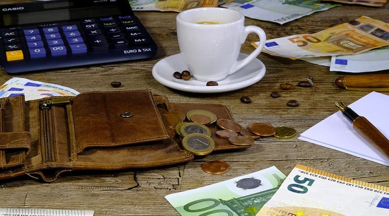 table en bois avec une calculatrice, un vieux portefeuille en cuir ouver, une tasse de café blanche, des billets de 50 et 100 euros avec des pieces.