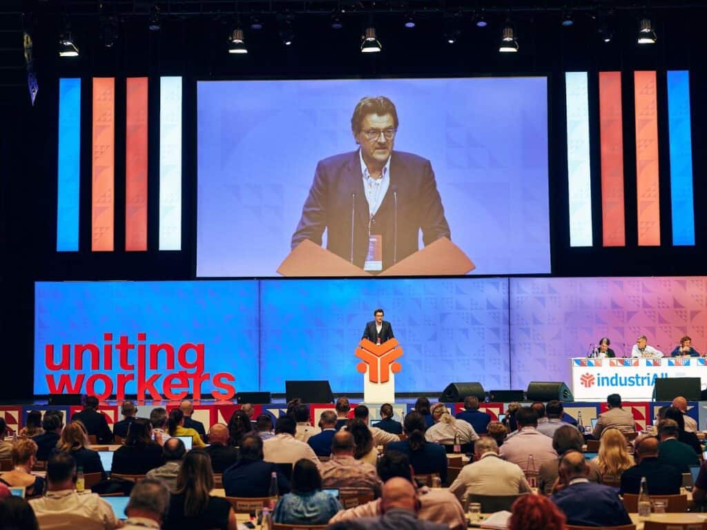 Salle de congres avec un homme à la tribune qui s'exprime, derriere lui sur grand ecran son visage en gros plan